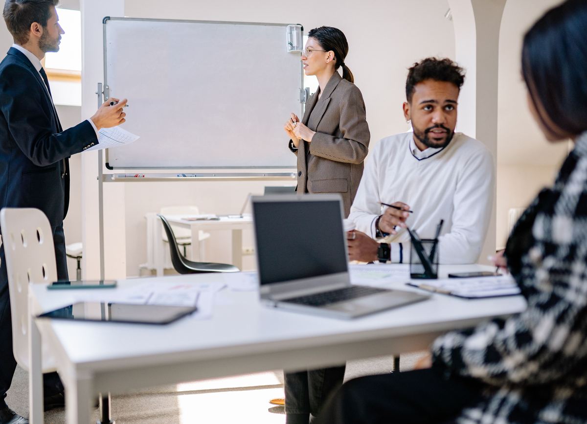 An operations meeting: one person at a whiteboard, colleagues at a table with a laptop and papers